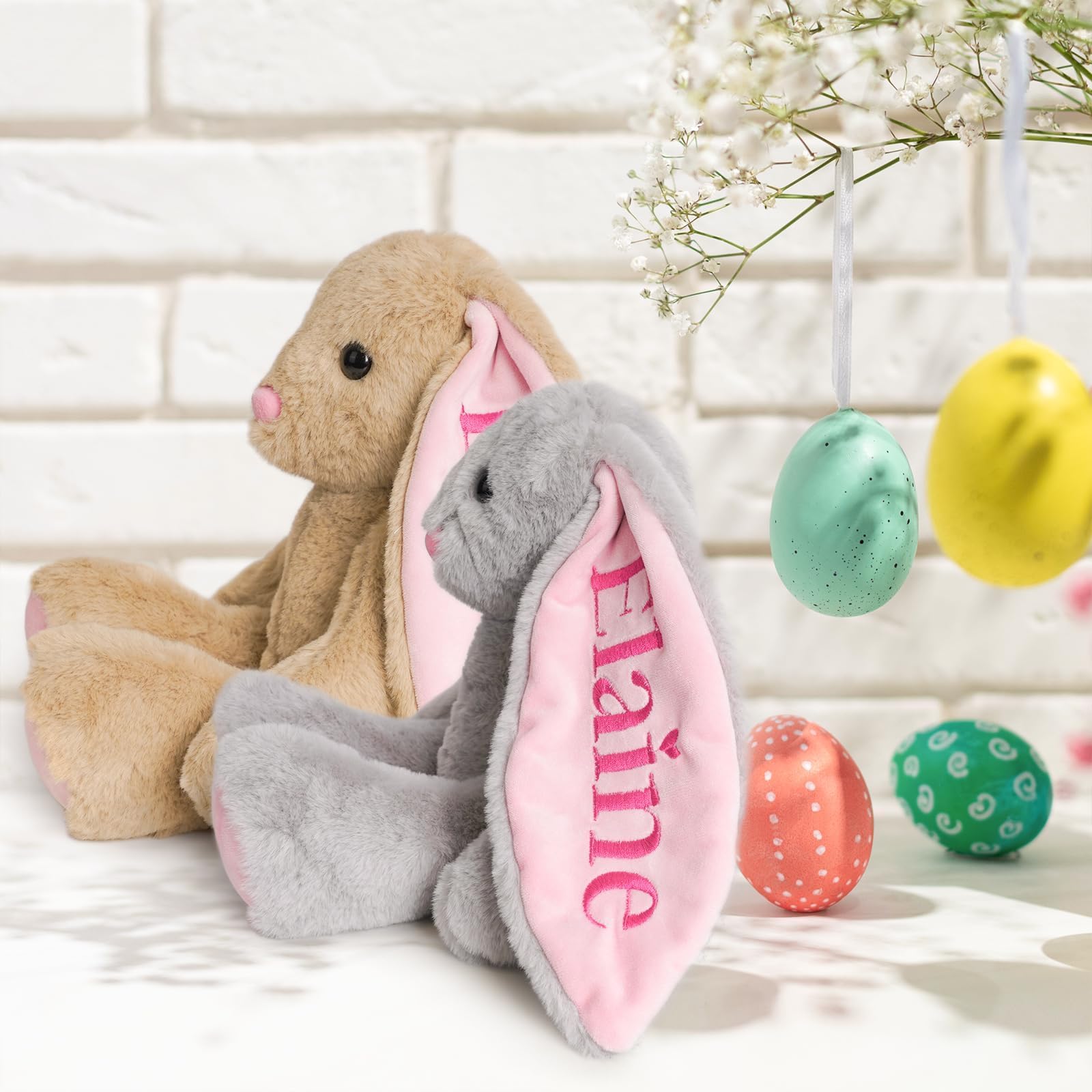 Personalized embroidered plush bunnies in grey and tan with names on pink ears, surrounded by colorful Easter eggs.